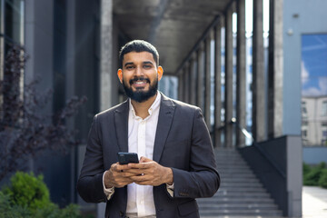 Wall Mural - Portrait of a Muslim male businessman standing on the street near a bank financial building and holding a phone in his hands, confidently and smilingly looking at the camera