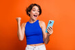 Photo of cheerful lucky girl dressed blue top rising fist winning game modern gadget isolated orange color background
