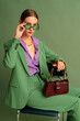  Fashionable beautiful confident woman wearing trendy color sunglasses, suit blazer, classic trousers, purple office shirt, chain necklace, holding brown leather bag, posing on green background