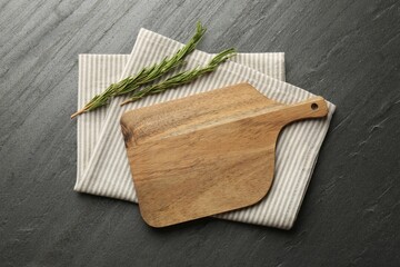 Wooden cutting board, kitchen towel and rosemary on dark grey table, top view