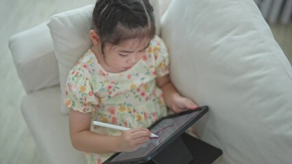 Wall Mural - Asian child girl using pen and touch drawing on tablet display screen. Baby smiling funny time to use tablet. Too much screen time. Cute girl watching videos while tv, Internet addiction concept.