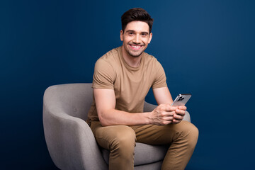 Sticker - Photo of toothy beaming intelligent man dressed beige t-shirt sit in armchair with smartpone in arms isolated on blue color background