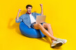 Full size photo of overjoyed guy wear jeans jacket shorts sit on pouf look at laptop win bet isolated on bright yellow color background