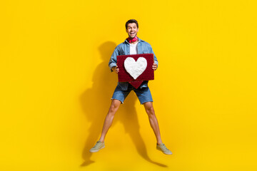 Poster - Full size photo of overjoyed guy wear jeans jacket shorts jumping hold heart tap like on comment isolated on yellow color background