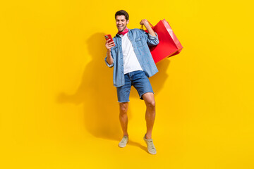 Poster - Full size photo of smart guy wear jeans jacket holding shopping bags read notification on smartphone isolated on yellow color background