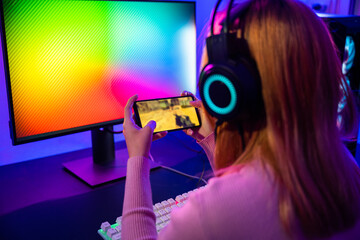 Wall Mural - Gamer playing online game application on mobile phone wear gaming headphones, Asian woman live stream she play video game via smartphone at home neon lights living room