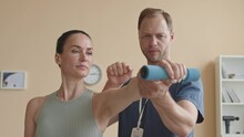 Tilt Up Shot Of Woman Holding Dumbbell And Bending Wrist While Male Physiotherapist Supporting Her Arm And Leading During Rehabilitation Exercise In ClinicTilt Up Shot Of Woman Holding Dumbbell And Be
