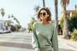 © Barbara Taylor - girl model wearing a plain bright sage green crewneck sweatshirt and jean shorts walking down a street with palm trees smiling with sunglasses