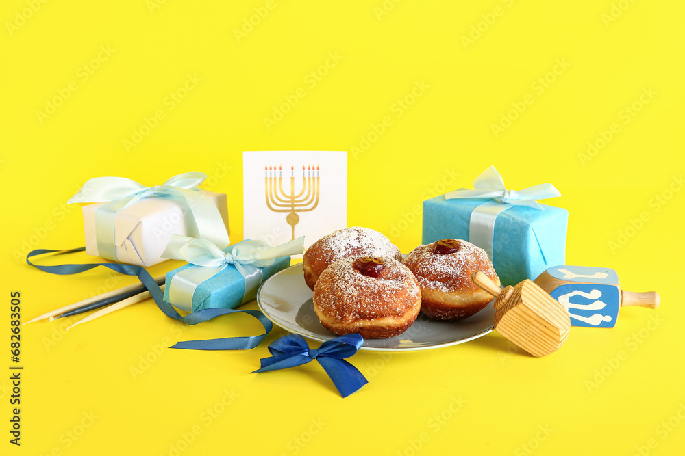 Dreidels with gift boxes, tasty donuts and greeting card on yellow background. Hanukkah celebration