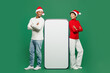 © ViDi Studio - Full body merry fun young couple man woman wear red casual clothes Santa hat posing big huge blank screen mobile cell phone isolated on plain green background. Happy New Year 2024 Christmas concept.