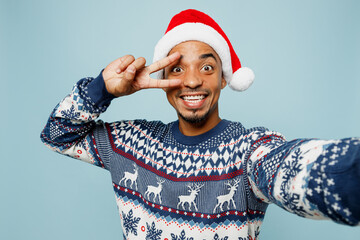 Wall Mural - Close up young man wearing sweater Santa hat posing doing selfie shot pov on mobile cell phone show v-sign isolated on plain blue background. Happy New Year 2024 celebration Christmas holiday concept.