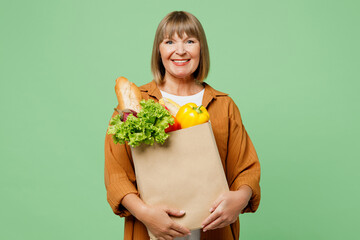 Elderly smiling happy cheerful positive woman wear brown shirt casual clothes hold shopping paper bag with food products looking camera isolated on plain green background. Delivery service from shop.