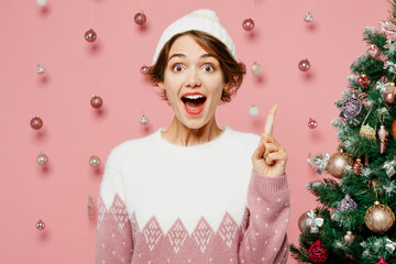 Wall Mural - Merry proactive young woman wears white sweater hat posing holding index finger up with great new idea isolated on plain pastel pink background. Happy New Year celebration Christmas holiday concept.