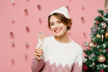 Wall Mural - Merry young woman wears white sweater hat posing hold in hand wineglass with champagne beverage isolated on plain pastel pink background studio. Happy New Year celebration Christmas holiday concept.