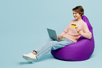 Wall Mural - Full body young smiling woman she wearing beige knitted sweater casual clothes sit in bag chair using mobile cell phone hold credit bank card doing online shopping isolated on plain blue background.