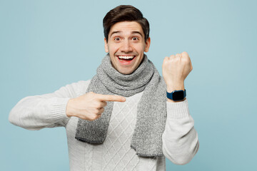 Young happy ill sick man wear gray sweater scarf show point on blank screen smart watch isolated on plain blue background studio Healthy lifestyle disease virus treatment cold season recovery concept