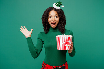 Wall Mural - Merry little kid teen girl wears hat casual clothes posing watch movie film hold bucket of popcorn in cinema isolated on plain green background. Happy New Year celebration Christmas holiday concept.