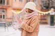 A young beautiful woman shakes snow off her knitted mittens against the backdrop of a winter snow-covered city.