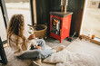© polinaloves - Young woman sitting by the fireplace in white sweater, drinking wine in cozy log cabin.