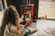 © polinaloves - Young woman sitting by the fireplace in white sweater, drinking wine in cozy log cabin.