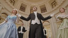 Low Angle Slowmo Of Ethnically Diverse Representatives Of Victorian Aristocracy Walking Along Pompous Baroque Ballroom With High Ornamental Ceiling And Golden Chandelier On It