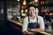  Asian handsome young male bartender standing with cross arm wearing descent suit with black apron and watch in the wrist looking in the camera with full bright smile