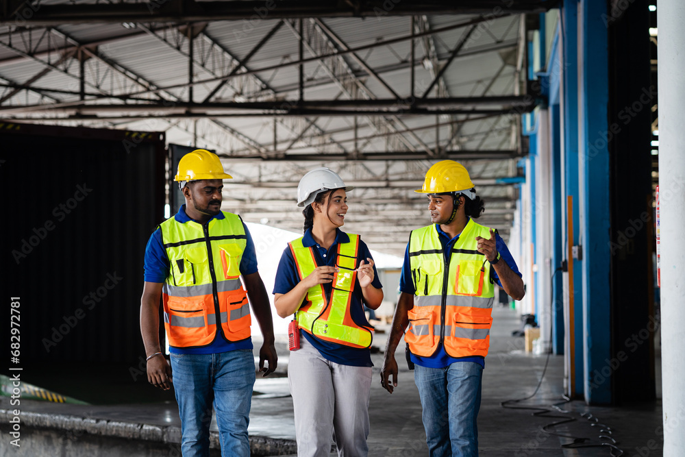 A team of warehouse workers in uniforms wearing yellow hard hats chats ...