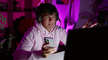 Poster - Attractive young hispanic man streamer immersed in digital technology, confidently smiling while having a lively video call on his smartphone in a dark gaming room, joyfully playing games