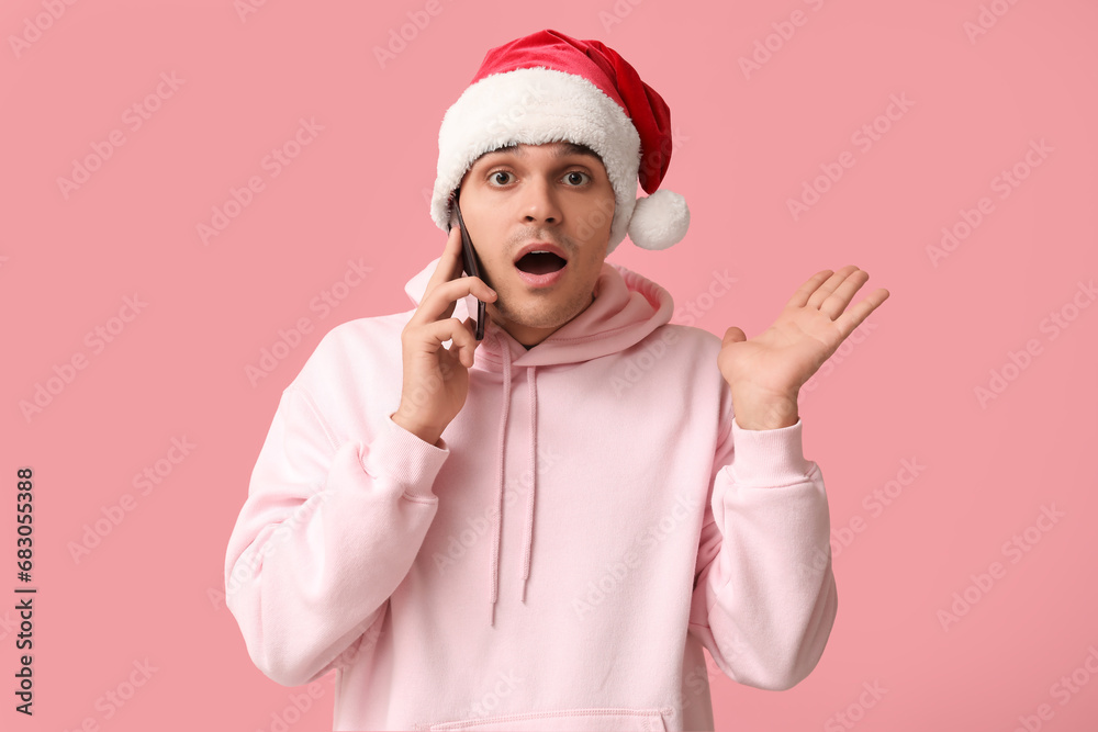 Shocked young man in Santa hat talking by phone on pink background