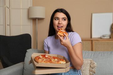 Canvas Print - Young woman eating piece of tasty pizza and holding cardboard box at home