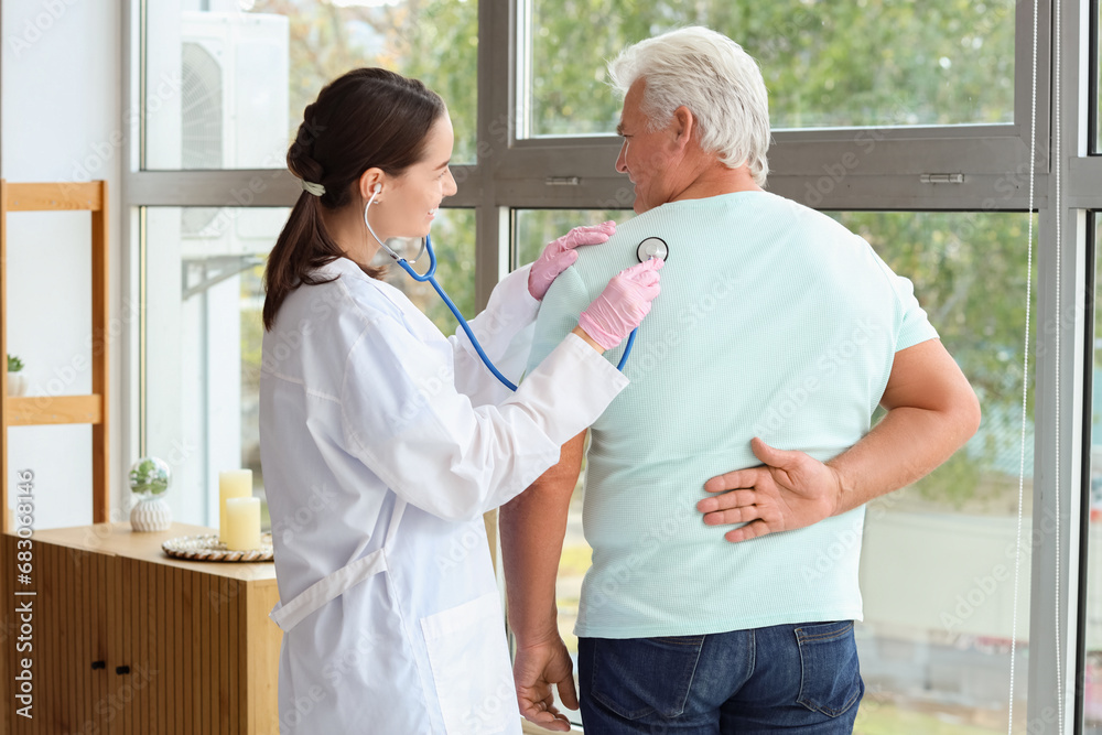 Nurse hearing senior man with stethoscope at home, back view