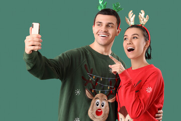 Sticker - Young couple in Christmas sweaters and reindeer horns taking selfie on green background