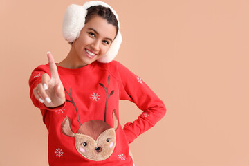 Canvas Print - Young woman in Christmas sweater and earmuffs pointing at something on beige background