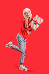 Canvas Print - Young woman in Santa hat and Christmas sweater with gift on red background