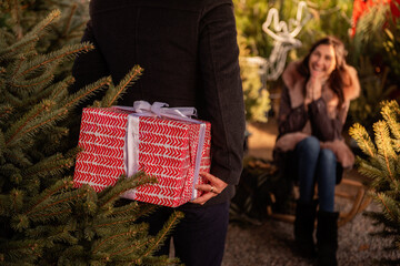 Young man holds large red box behind his back. Beautiful woman covers her face with hands. Couple in love among green Christmas tree market. Boxing Day. Showing attention during holidays. Lifestyle