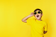 Excited young boy in vibrant yellow lifting sunglasses on a matching background, expressing surprise and joy.