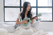 © kaew6566 - Cheerful asian woman playing ukulele sitting on white bed with pillows beside window at apartment. Joyful young girl holding acoustic ukulele in morning at home. Relax female play music ukulele hobby.