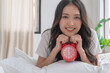 © kaew6566 - Beautiful cheerful asian woman early wake up lying relax on pillow holding hand red alarm clock at home. Happy young girl refreshing early wake up on white with alarm clock looking camera in morning.