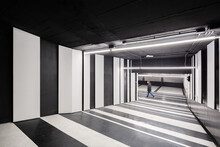 Pedestrian Walking Through A Modern Parking Garage With Striking Black And White Striped Walls And Floor Patterns, Illuminated By Overhead Fluorescent Lights