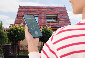 Canvas Print - Woman using smart home control system via mobile phone near house outdoors, closeup