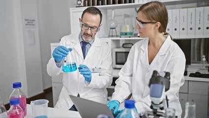 Canvas Print - Two jubilant scientists working together in a lab, conversing and using a laptop while holding a test tube for sample analysis in their indoor science research workplace.