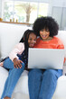 Happy african american mother and daughter siyying on couch and using laptop at home, copy space