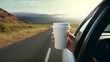 hand with a white paper coffee cup stretched out of the window of a car driving in nature.