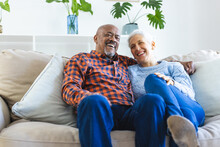 Happy Diverse Senior Couple Sitting On Sofa And Embracing In Sunny Living Room
