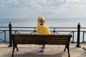 Wall Mural - A young girl in a yellow T-shirt sits on a bench by the sea and admires the seascape in Athens.