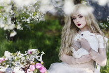 Beautiful Girl With A White Rabbit In A Blooming Apple Garden. White Flowers In The Garden In Sunlight. Spring Apple Trees In Bloom. Beautiful Girl Among Spring Blossoms