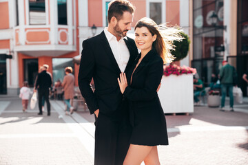 Beautiful fashion woman and her handsome elegant boyfriend in black suit. Sexy brunette model in jacket. Fashionable confident couple posing in street. Brutal man and female outdoors