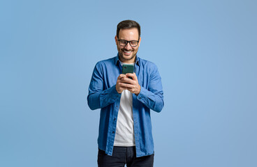 Wall Mural - Male freelancer smiling and using social media over smart phone while standing on blue background