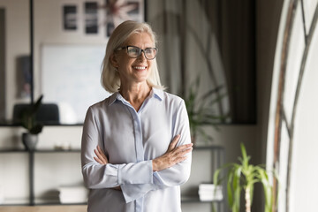 Canvas Print - Happy pensive elder business leader woman office portrait. Confident senior businesswoman in elegant glasses, mature professional with arms crossed looking away with toothy smile