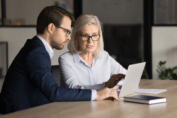 Focused senior business professional complaining on laptop, software work problem to application developer, computer expert, sitting at laptop, pointing at display, speaking